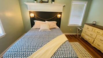A bedroom with a patterned bedspread, a headboard with wall-mounted lights, a dresser, and a window with blinds.