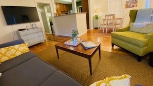 Un salon avec un canapé, une table basse et un téléviseur. On aperçoit un coin cuisine au fond.