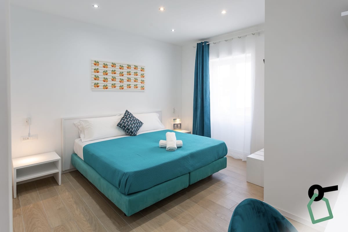 A modern bedroom with a teal bedspread, a white headboard, a wall-mounted fruit-themed artwork, a bedside table with a lamp, and a window with blue curtains.
