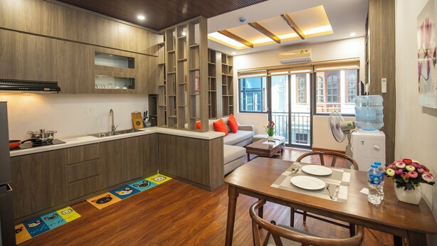 A modern kitchen with wooden cabinets, a built-in stove, and a dining area with a table set for two. - Sumitomo7- Linh Lang Serviced Apartment (Hanoi)
