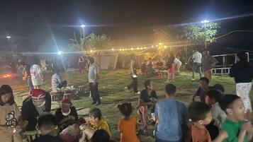 An outdoor gathering with people seated and standing, under string lights, on a grassy area.