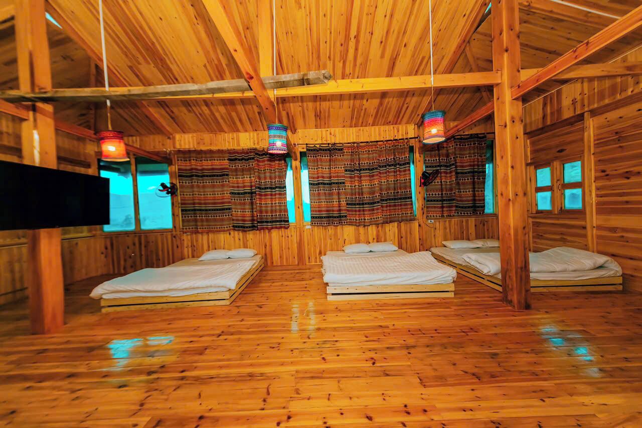 Una cabaña con cuatro camas de madera dispuestas en fila, cada una con almohada y manta. Hay tres lámparas colgantes con tonalidades coloridas.