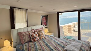 A bedroom with a large bed, a nightstand, a lamp, a framed picture, and a view of the sea through a balcony. - Stay with the best view of Izmir (İzmir)
