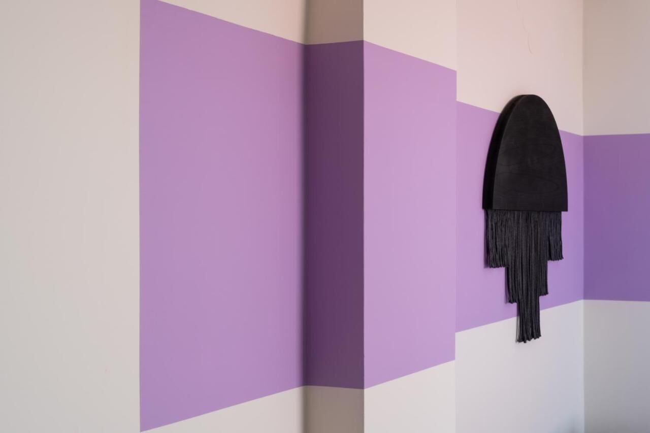 Una habitación con una decoración morada y blanca, un objeto negro colgado en la pared y una vista parcial de una puerta.