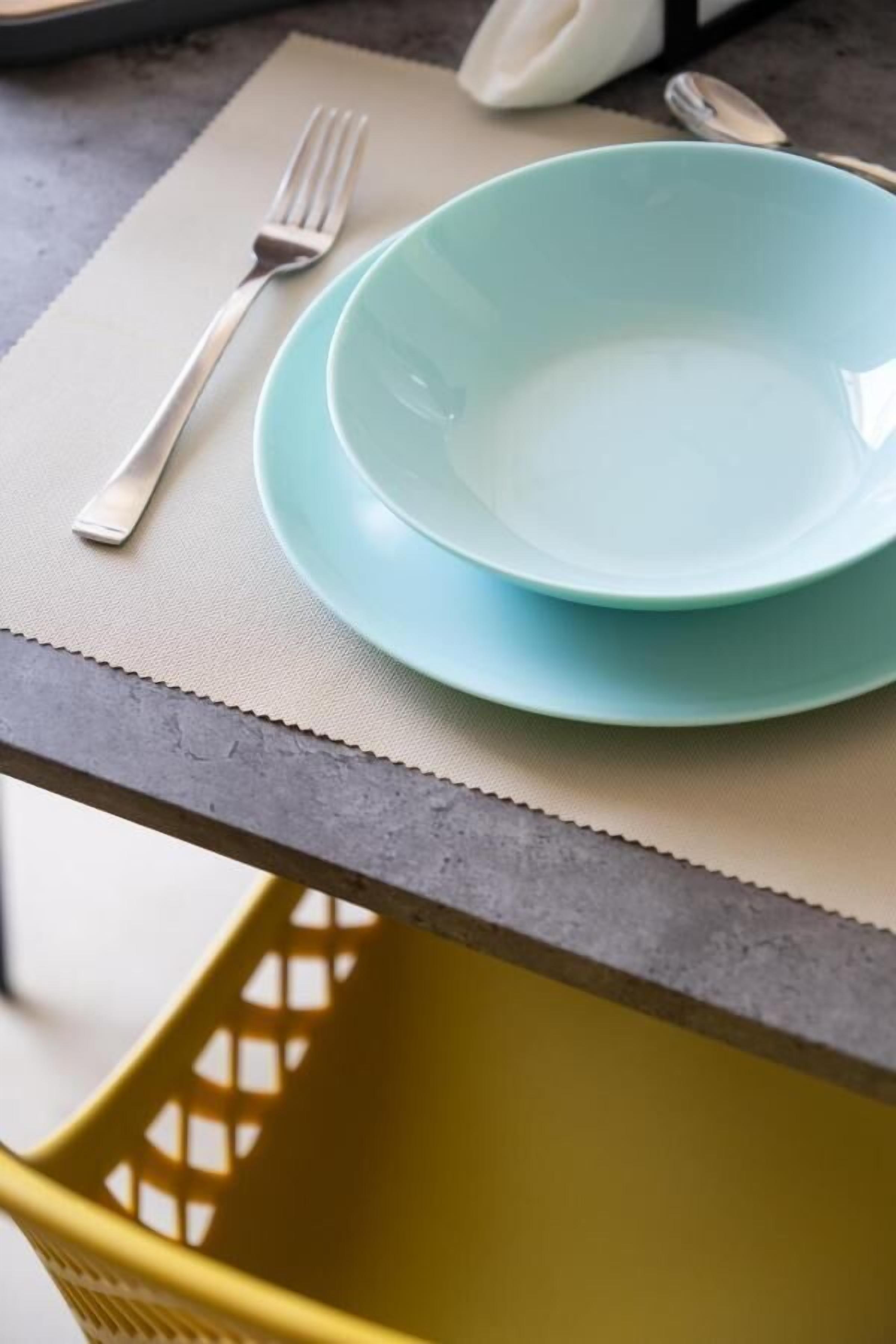 A dining setup with a light blue bowl and plate on a beige placemat, a fork, and a spoon on the table.