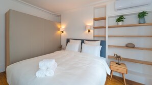 Un lit bien fait, avec du linge de lit blanc et des serviettes pliées, une table de chevet avec une lampe et une étagère murale agrémentée d’objets décoratifs.