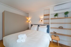 Una cama bien tendida con sábanas blancas y toallas enrolladas, una mesita de noche con lámpara, y un estante en la pared con objetos decorativos.