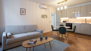 A modern living room with a sofa, coffee table, and chairs. A kitchen with cabinets and a dining area is visible in the background.