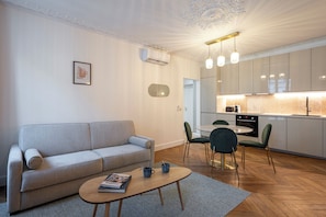 A modern living room with a sofa, coffee table, and chairs. A kitchen with cabinets and a dining area is visible in the background.