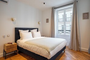 A neatly made bed with white linens in a room with a wooden floor, a small bedside table, and a window with curtains. - Madeleine Opéra - Excellent One Bedroom in Paris (Paris)
