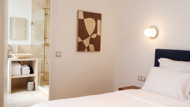A hotel room with a bed, a bedside table, a wall-mounted light, a bathroom with a sink and shower, and a towel rack.