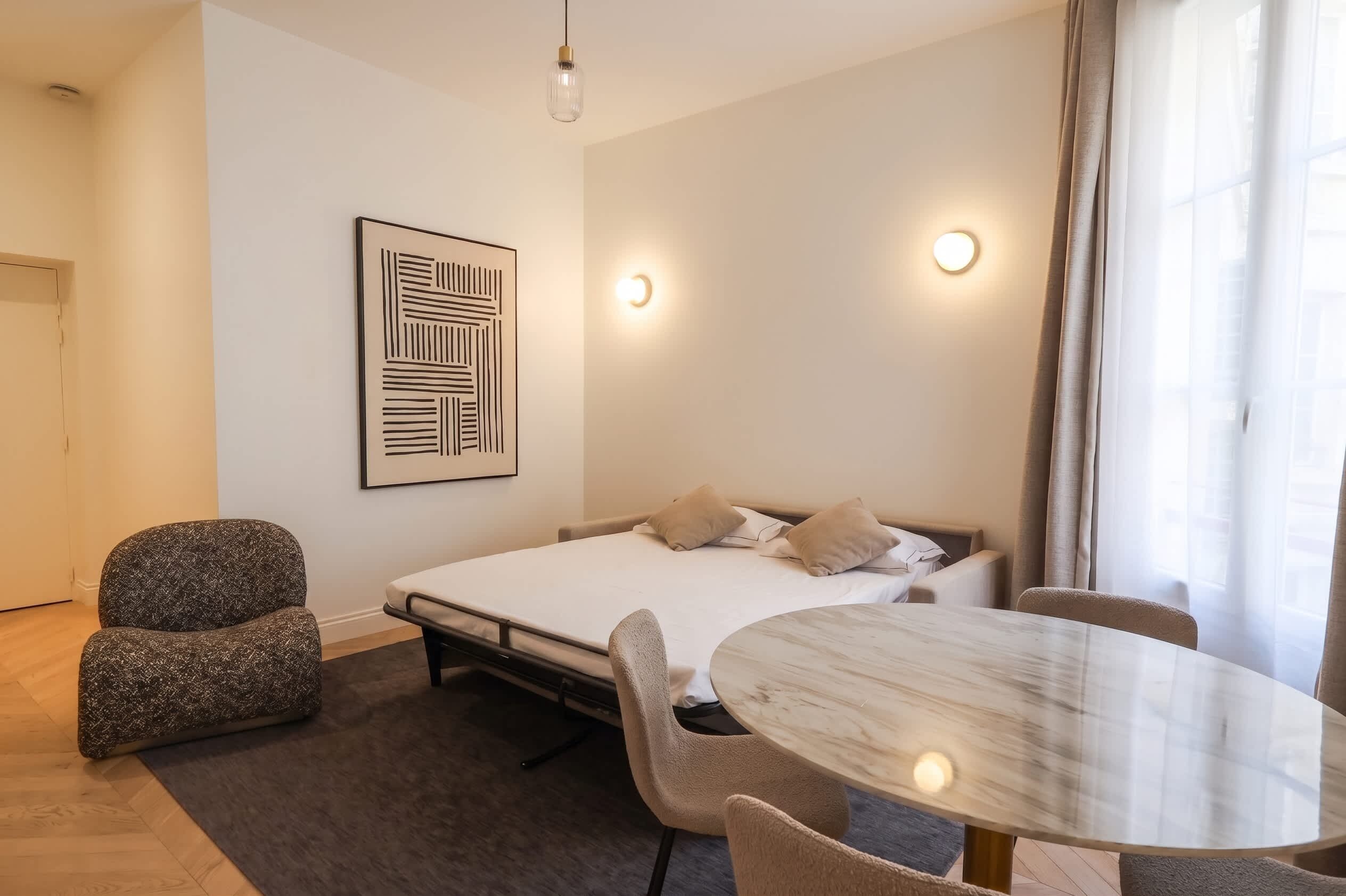 Une chambre à coucher moderne avec un lit, une chaise, une table ronde et un luminaire fixé au mur.