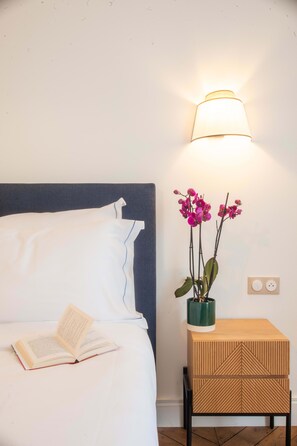 A neatly made bed with a pillow and an open book, a bedside lamp, a small wooden nightstand with a geometric pattern, and a vase with pink orchids.