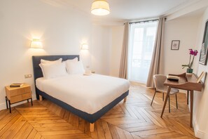 A neatly arranged hotel room with a large bed, bedside lamps, a wooden herringbone floor, a desk with a chair, and a window with curtains. - Madeleine Opéra - Excellent One Bedroom in Paris (Paris)