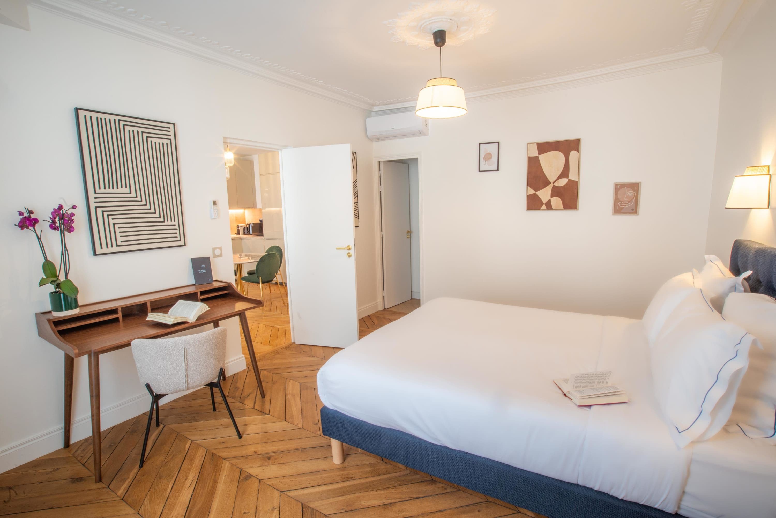 Ein modernes Hotelzimmer mit Holzboden im Fischgrätmuster, einem großen Bett mit weißer Bettwäsche, einem Schreibtisch mit Stuhl und einem kleinen Tisch mit Blumen.