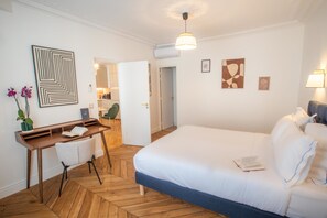 A modern hotel room with a wooden herringbone floor, a large bed with white linens, a desk with a chair, and a small table with a vase of flowers. - Madeleine Opéra - Excellent One Bedroom in Paris (Paris)