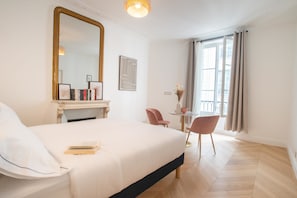Quarto de hotel com uma cama grande, lareira, mesa pequena com cadeiras e uma janela ampla com cortinas.