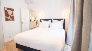 A hotel room with a bed, bedside tables, a nightstand, a wall-mounted air conditioner, a door leading to a bathroom, and a kitchen area visible through an open door. - Madeleine Opéra - Excellent One Bedroom in Paris (Paris)