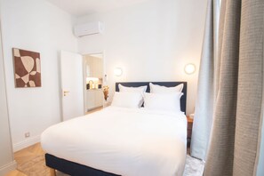 A hotel room with a bed, bedside tables, a nightstand, a wall-mounted air conditioner, a door leading to a bathroom, and a kitchen area visible through an open door. - Madeleine Opéra - Excellent One Bedroom in Paris (Paris)