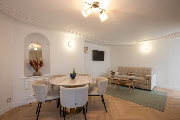 A modern living room with a round dining table, four chairs, a sofa, a flat-screen TV, and a decorative vase.