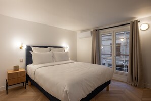 A neatly arranged bedroom with a large bed, bedside lamps, a small wooden nightstand, and a window with curtains.