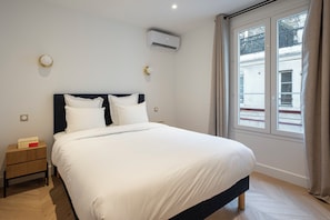 A neatly made bed with white linens, a bedside table with a geometric-patterned nightstand, and a window with curtains. - Madeleine Opéra - Fabulous Two Bedroom in Paris (Paris)