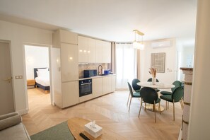A modern kitchen with white cabinets, a dining area with a round table and green chairs, and a visible bed in the adjacent room.