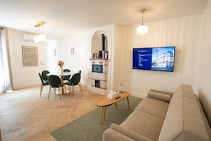 A modern living room with a sofa, a coffee table, a fireplace, a dining area with chairs, and a mounted TV displaying a list. - Madeleine Opéra - Fabulous Two Bedroom in Paris (Paris)