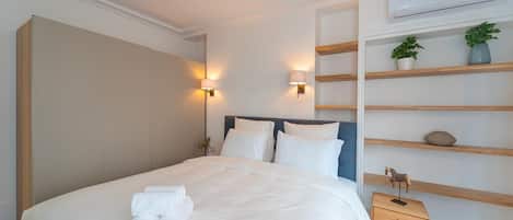 Una cama bien tendida con sábanas blancas y toallas enrolladas, una mesita de noche con lámpara, y un estante en la pared con objetos decorativos.