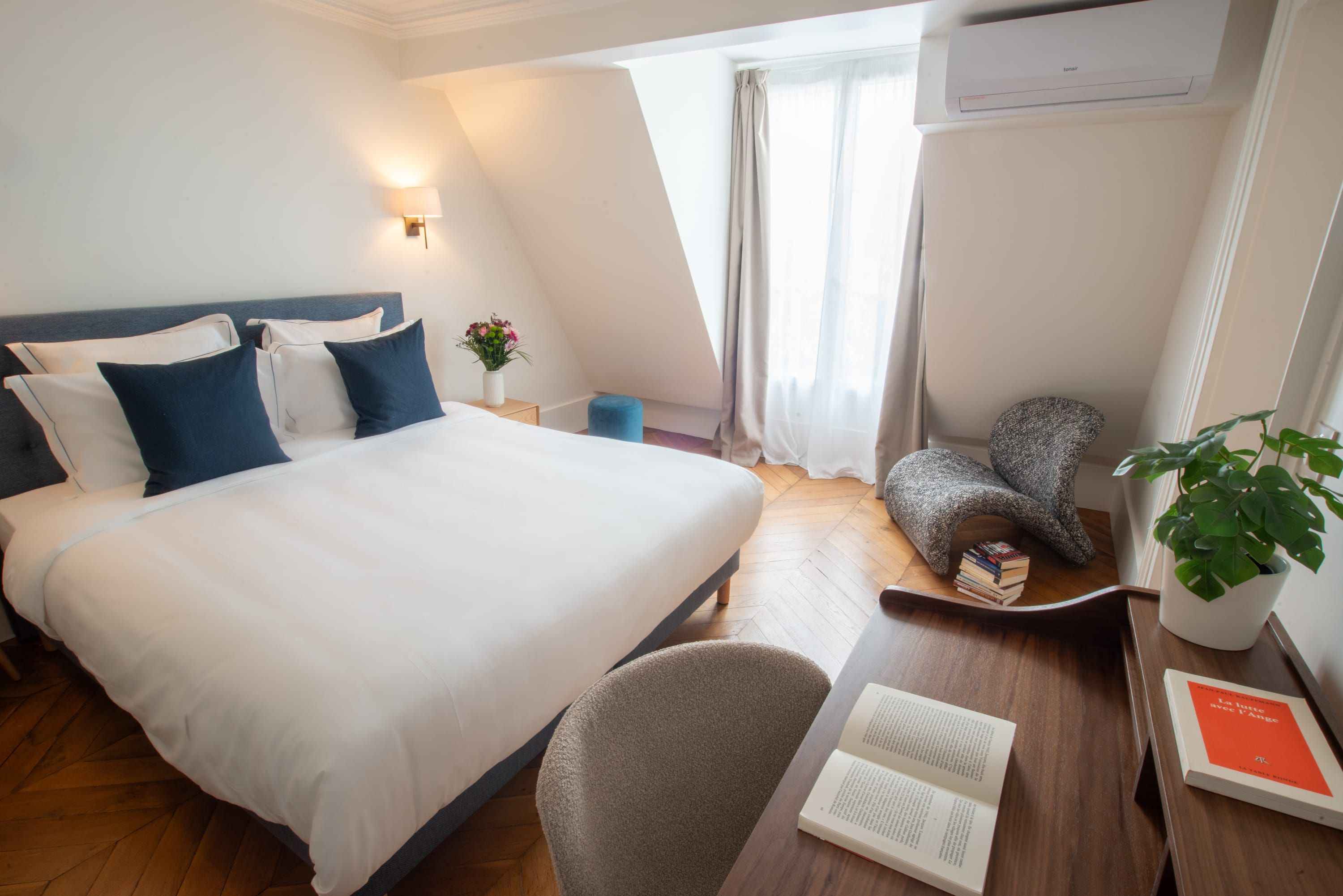 Une chambre d’hôtel avec un grand lit, un bureau avec des livres, une chaise, une petite table avec une plante et une fenêtre avec des rideaux.
