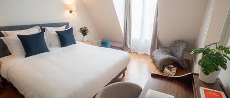 Habitación de hotel con una cama grande, un escritorio con libros, una silla, una mesita con una planta y una ventana con cortinas.