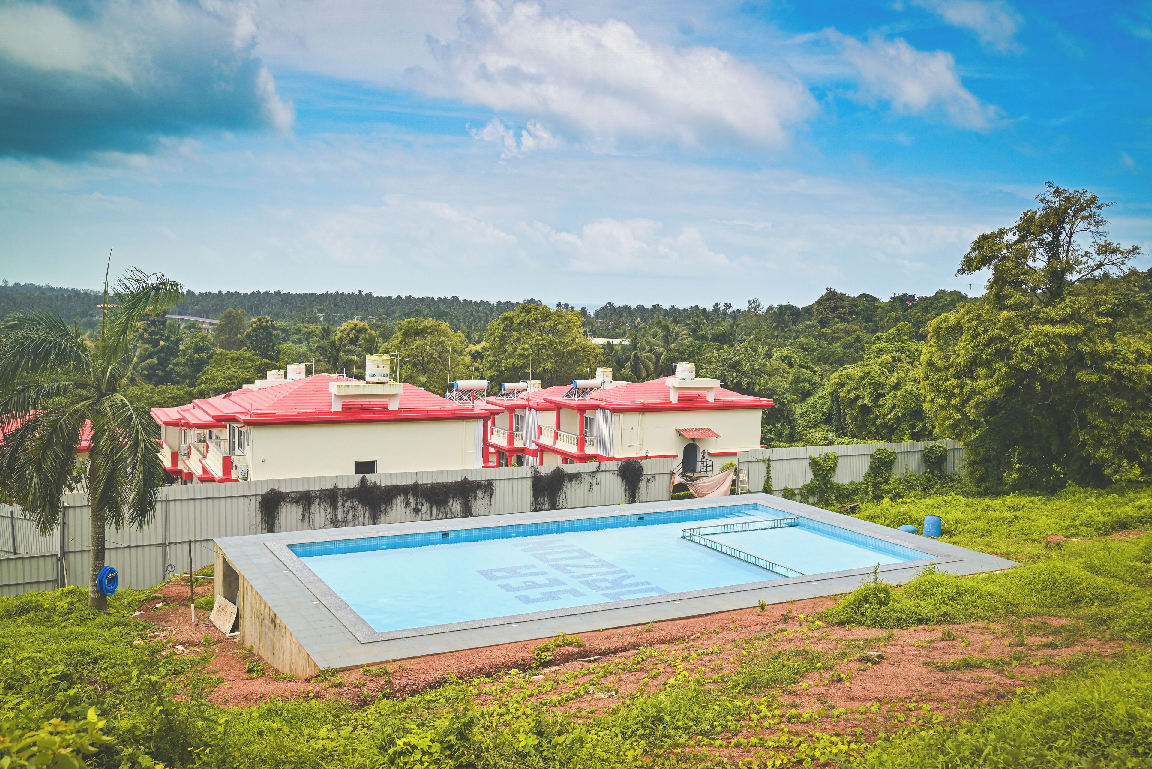 A swimming pool surrounded by a fenced area with red-roofed buildings in the background.