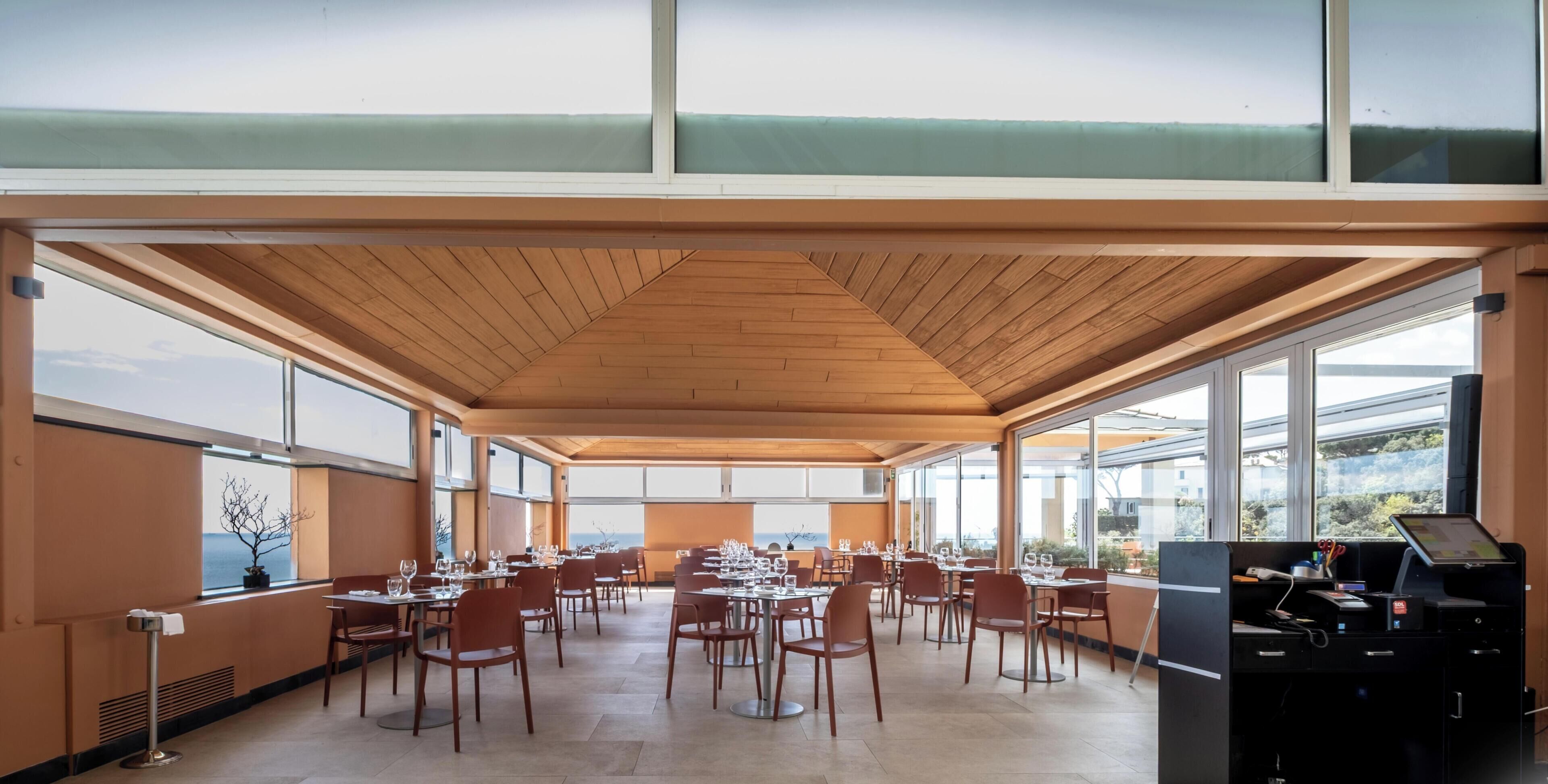 Un comedor amplio con varias mesas y sillas, ventanas grandes que ofrecen vistas y un techo de madera.