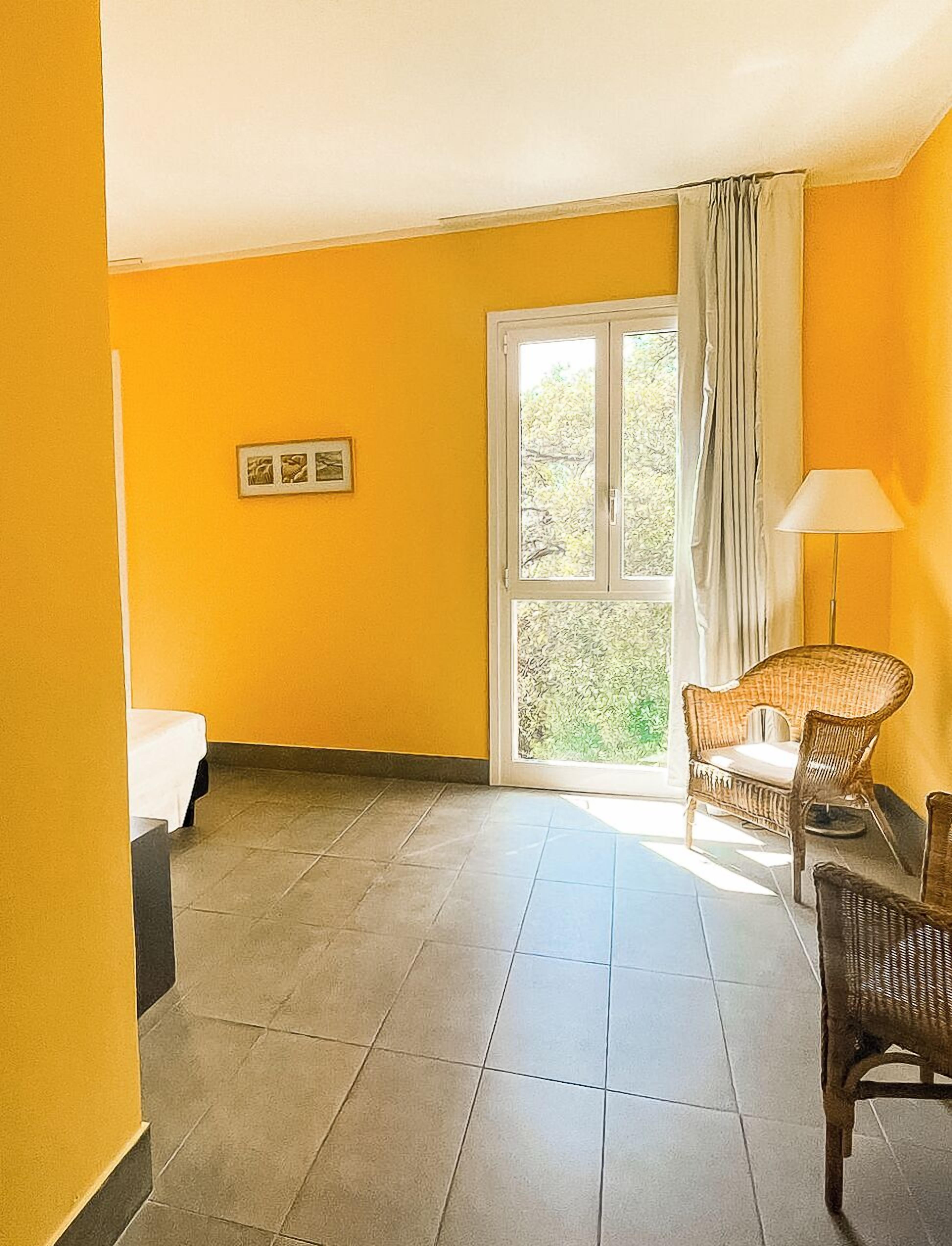 Une pièce aux murs jaunes, avec un lit, une fenêtre avec des rideaux et des chaises en osier.