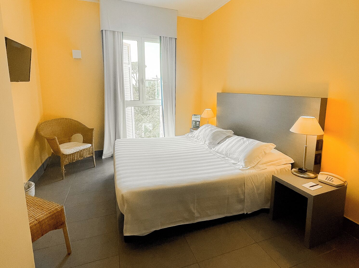 Habitación de hotel con una cama grande, una silla, una mesita de noche con lámpara y una ventana con cortinas.