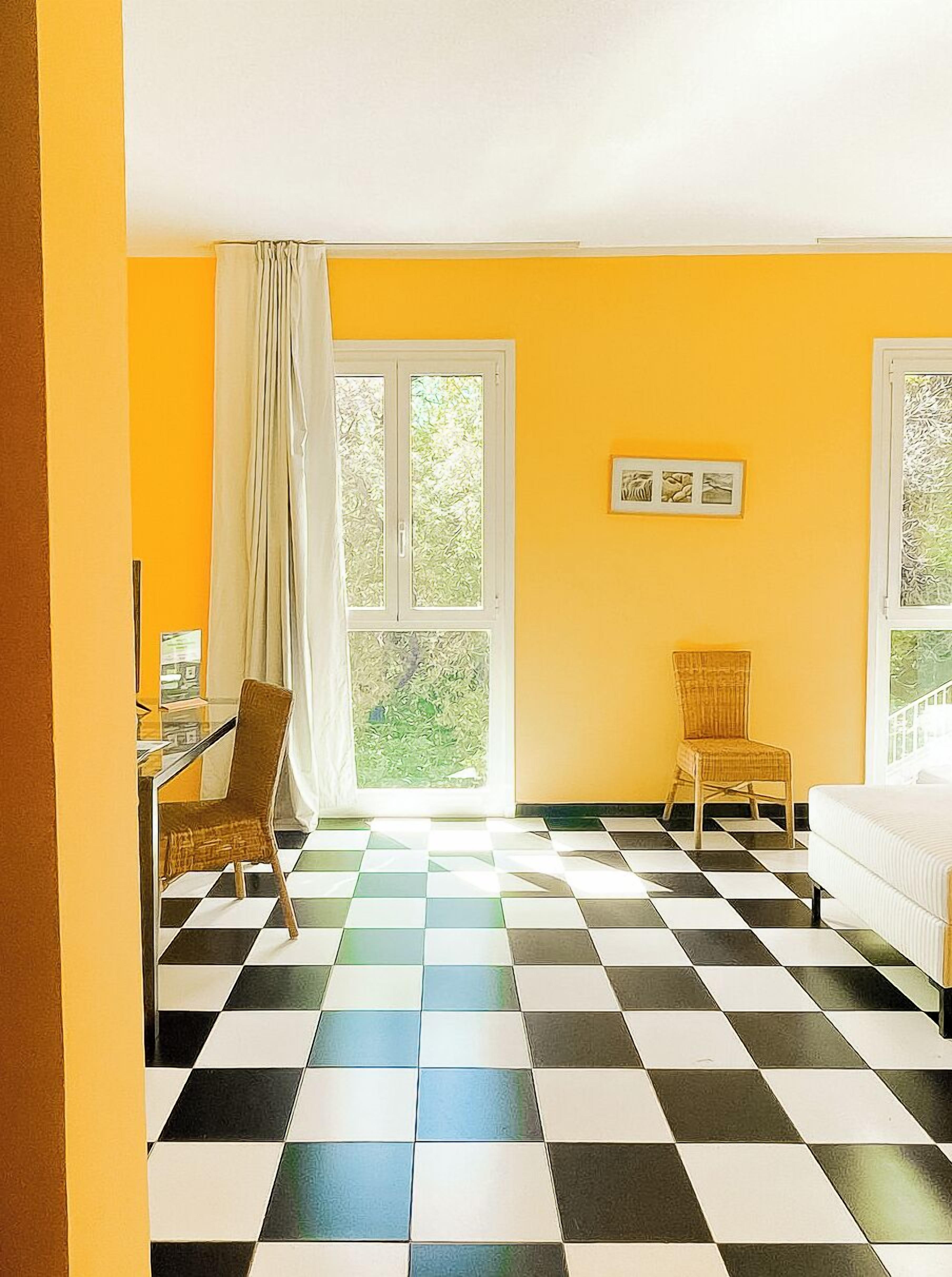 A room with a checkered floor, a yellow wall, and a window with a view of greenery.