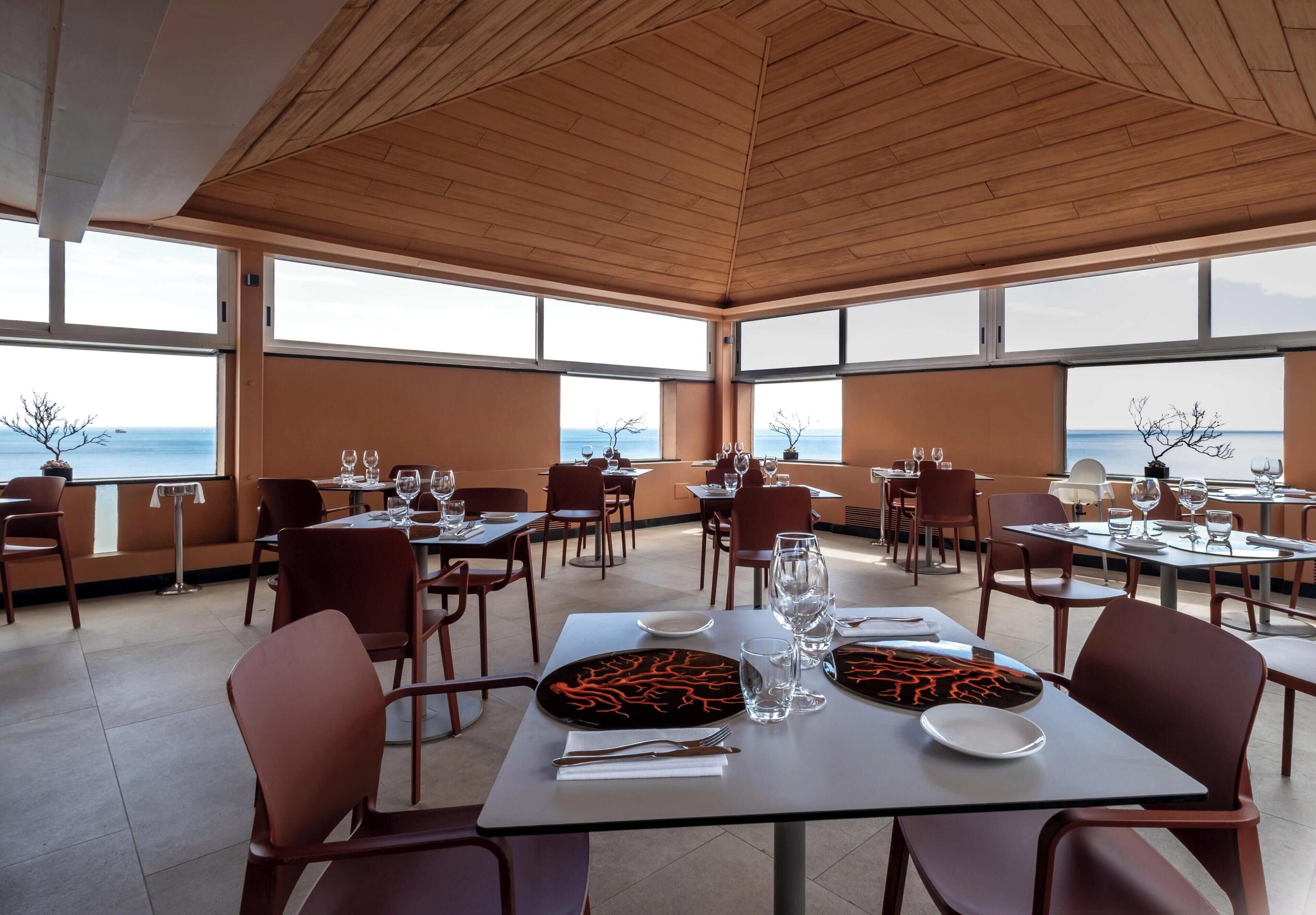 An indoor dining area with tables and chairs, large windows, and a view of the sea.