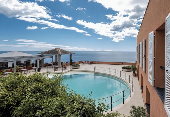 A pool area with a view of the sea, a covered seating area, and a building with shutters. - Hotiday Arenzano Pineta (Arenzano)