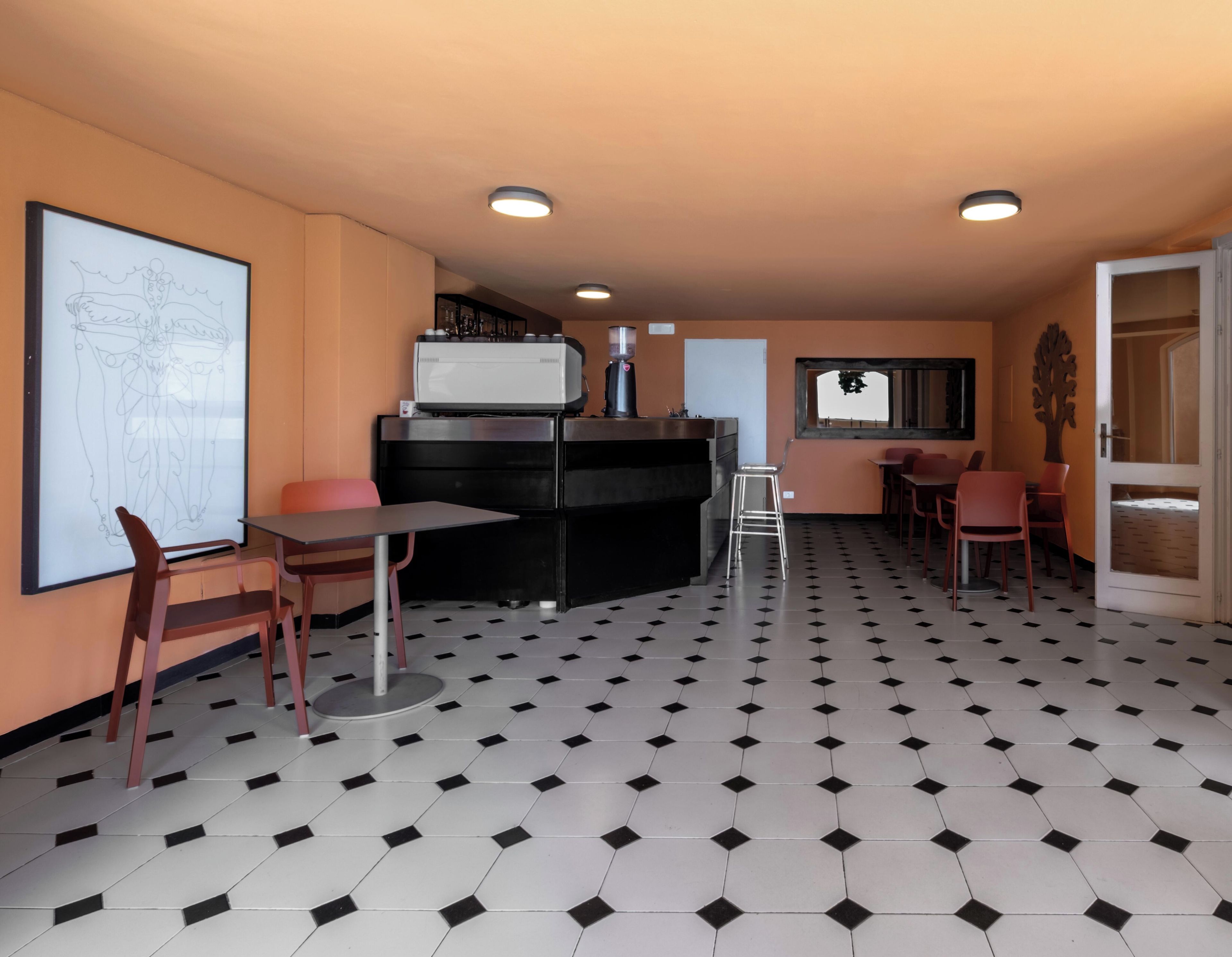 A room with a black and white checkered floor, a whiteboard with a drawing, a table with chairs, a coffee machine, and a door with a mirror.
