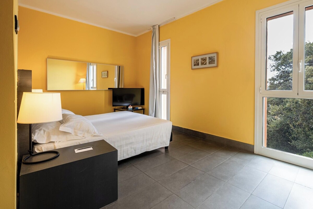 A hotel room with a bed, a TV, a desk with a lamp, and a window with a view of trees.