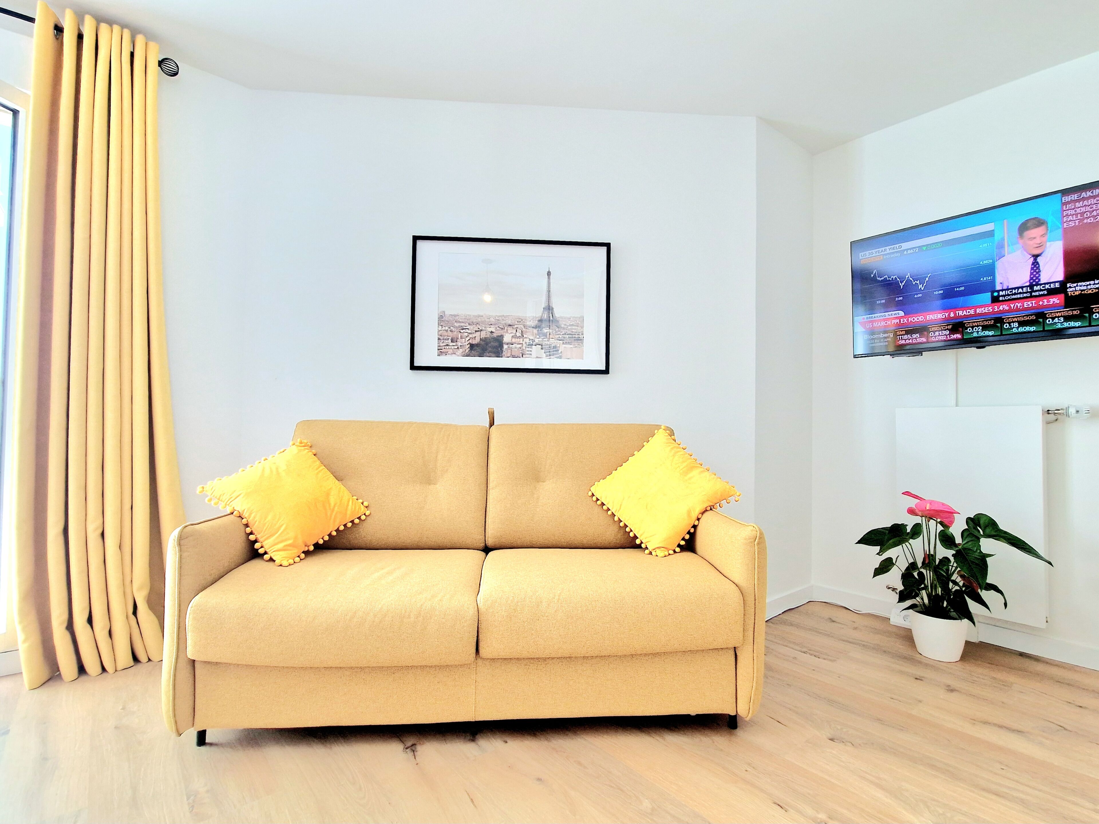 A living room with a beige sofa, two yellow pillows, a framed cityscape picture, a television displaying financial news, and a potted plant.