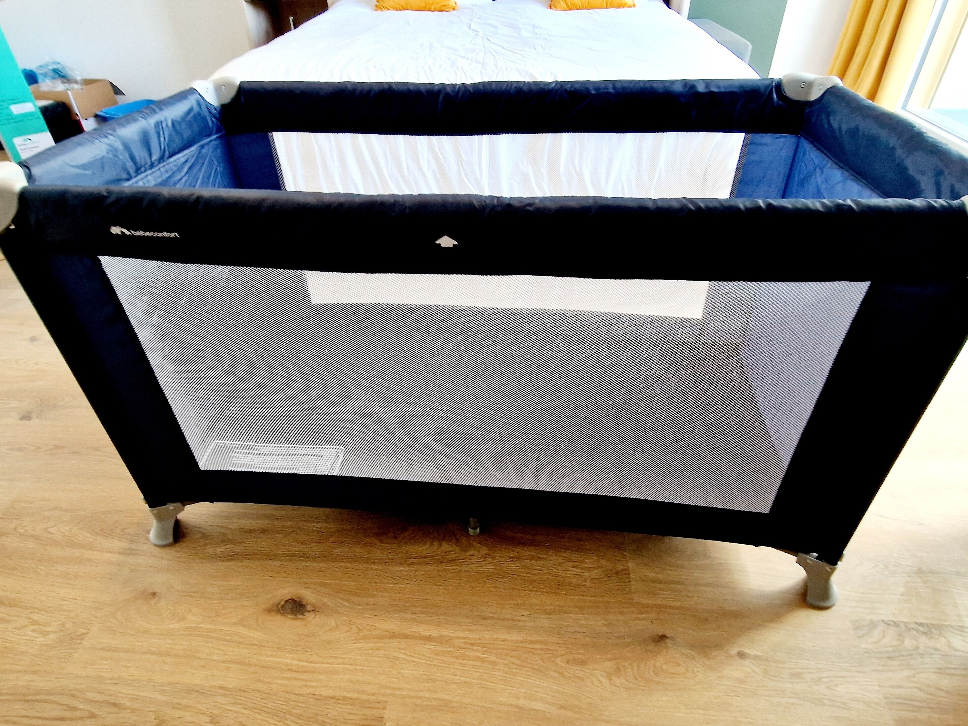 A portable crib with a mesh side and a white mattress, placed on a wooden floor.