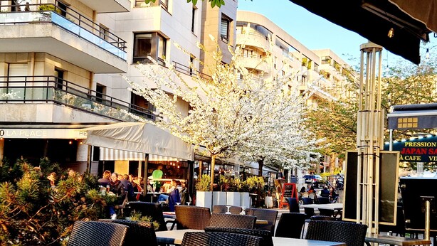 An outdoor cafe with tables and chairs, surrounded by buildings and a tree with white blossoms. - Stylish Stay Near Paris - 850m to Subway RER A - Netflix 1BR-4P (Rueil-Malmaison)