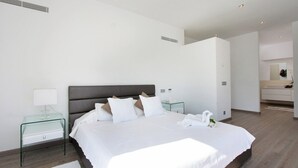 Una habitación de hotel moderna con una cama grande, una mesita de vidrio, una lámpara de noche y un baño visible a través de una puerta abierta.
