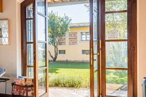 Une pièce dotée d’une grande porte vitrée donnant sur un jardin, d’une petite table avec des livres, et offrant une vue sur un bâtiment dont le toit est recouvert de tuiles.