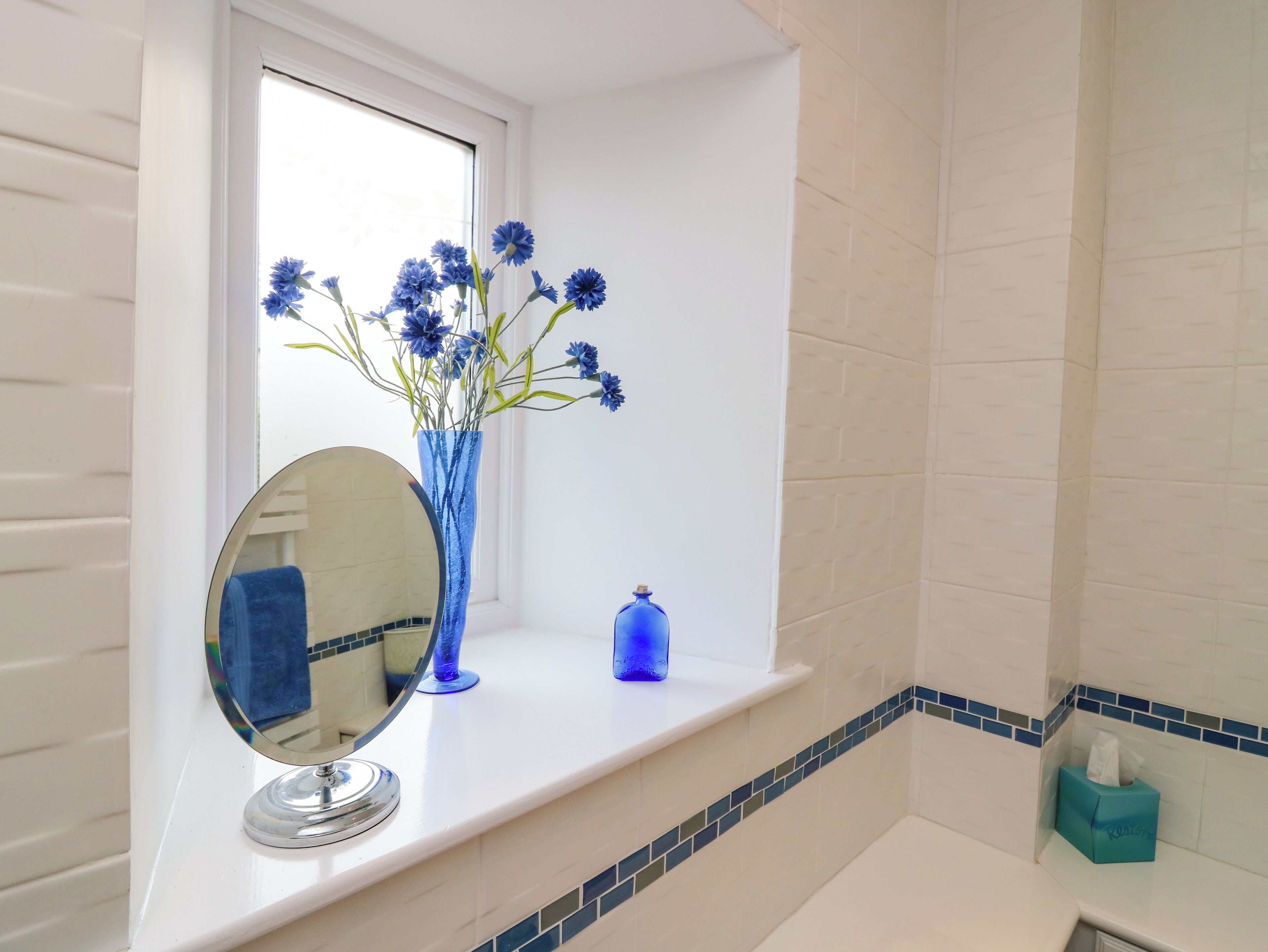 A bathroom with a window, a mirror, a vase with flowers, and a blue soap dispenser.