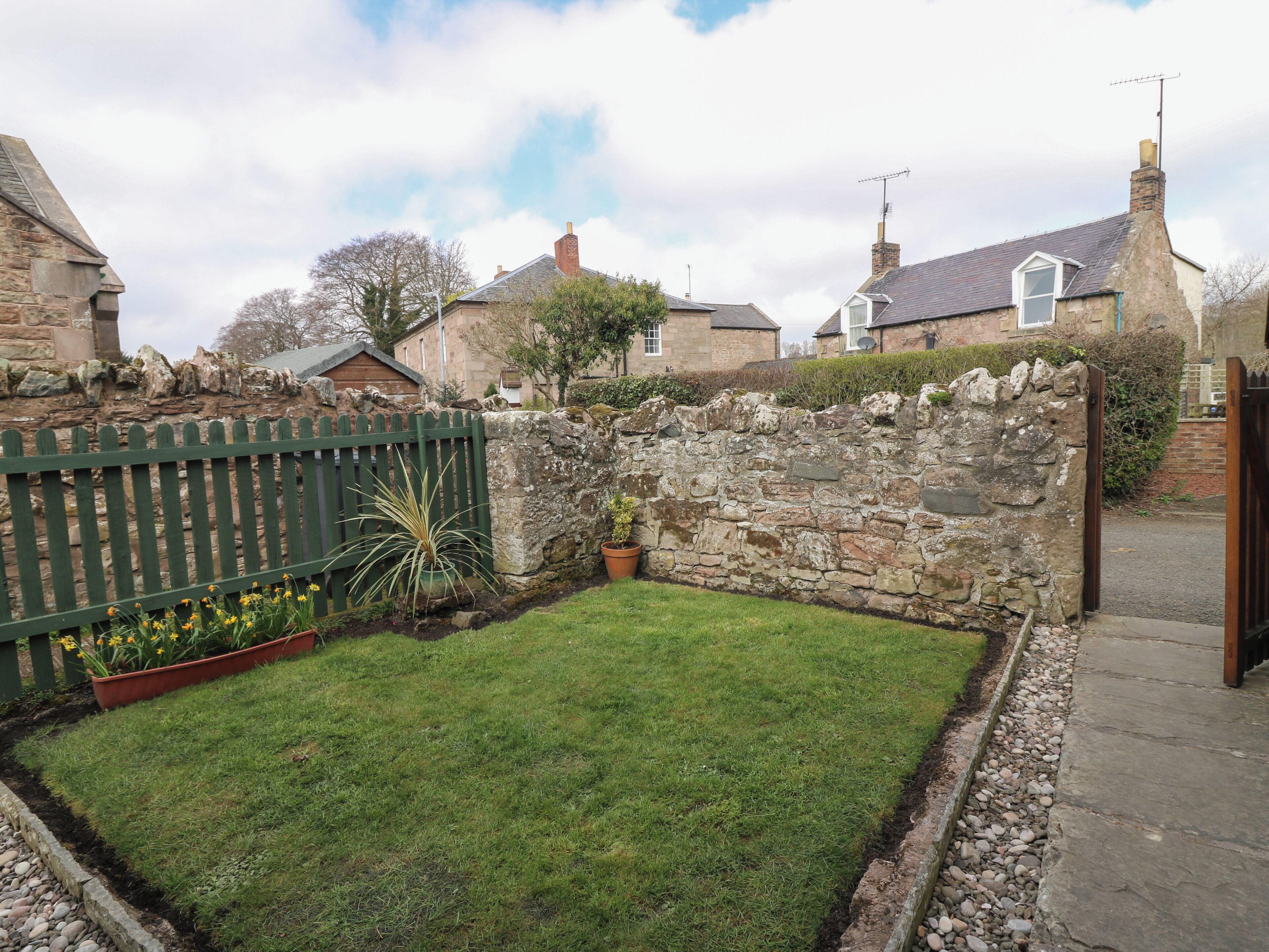 A small, well-maintained garden with a stone wall, a wooden fence, and a paved path leading to a gate.