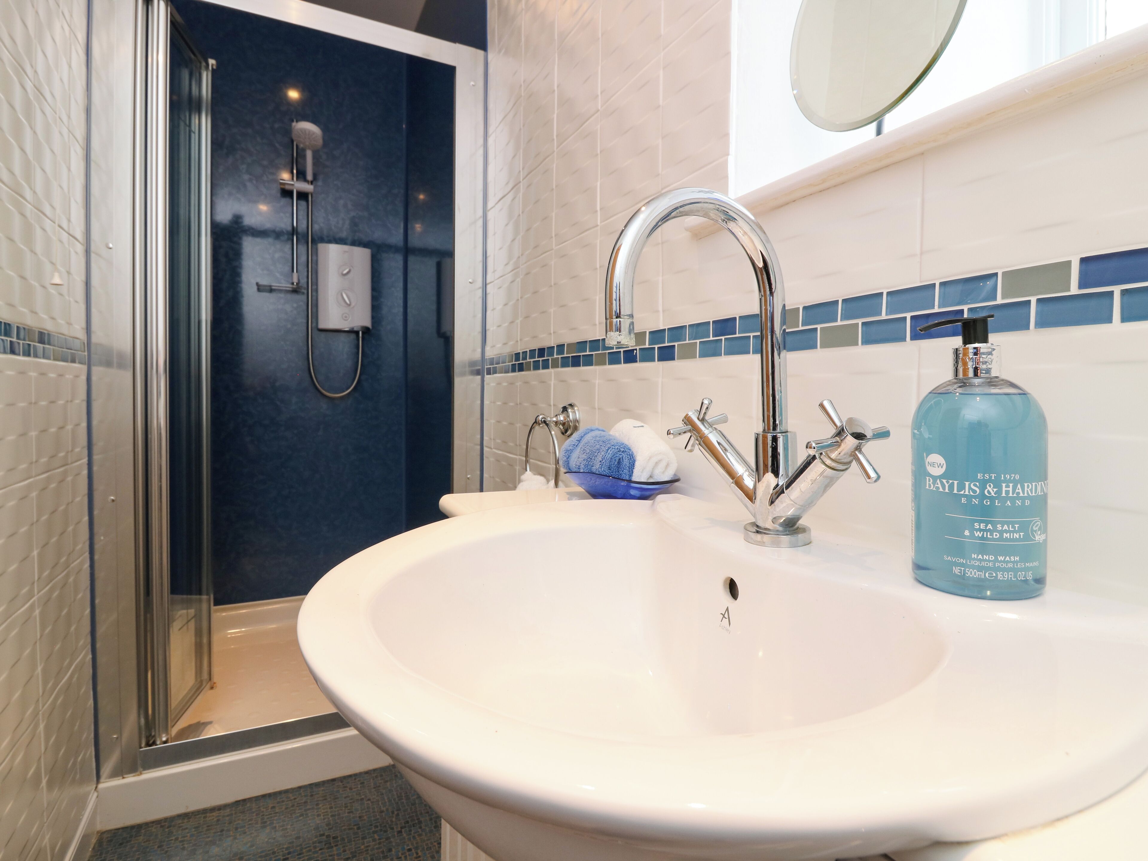 A bathroom with a white sink, a handheld showerhead, and a blue bottle of Baylis & Hardin hand wash.