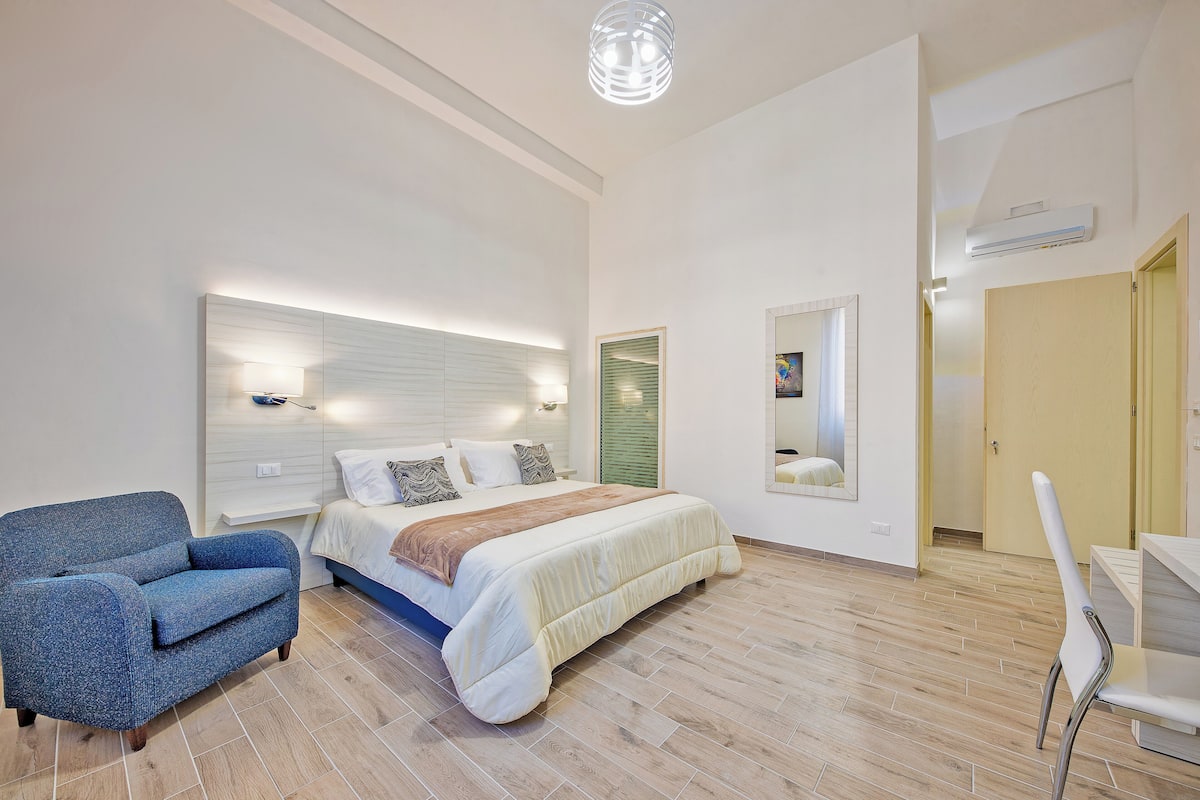 Quarto moderno com cama, poltrona azul, mesa de cabeceira e espelho.