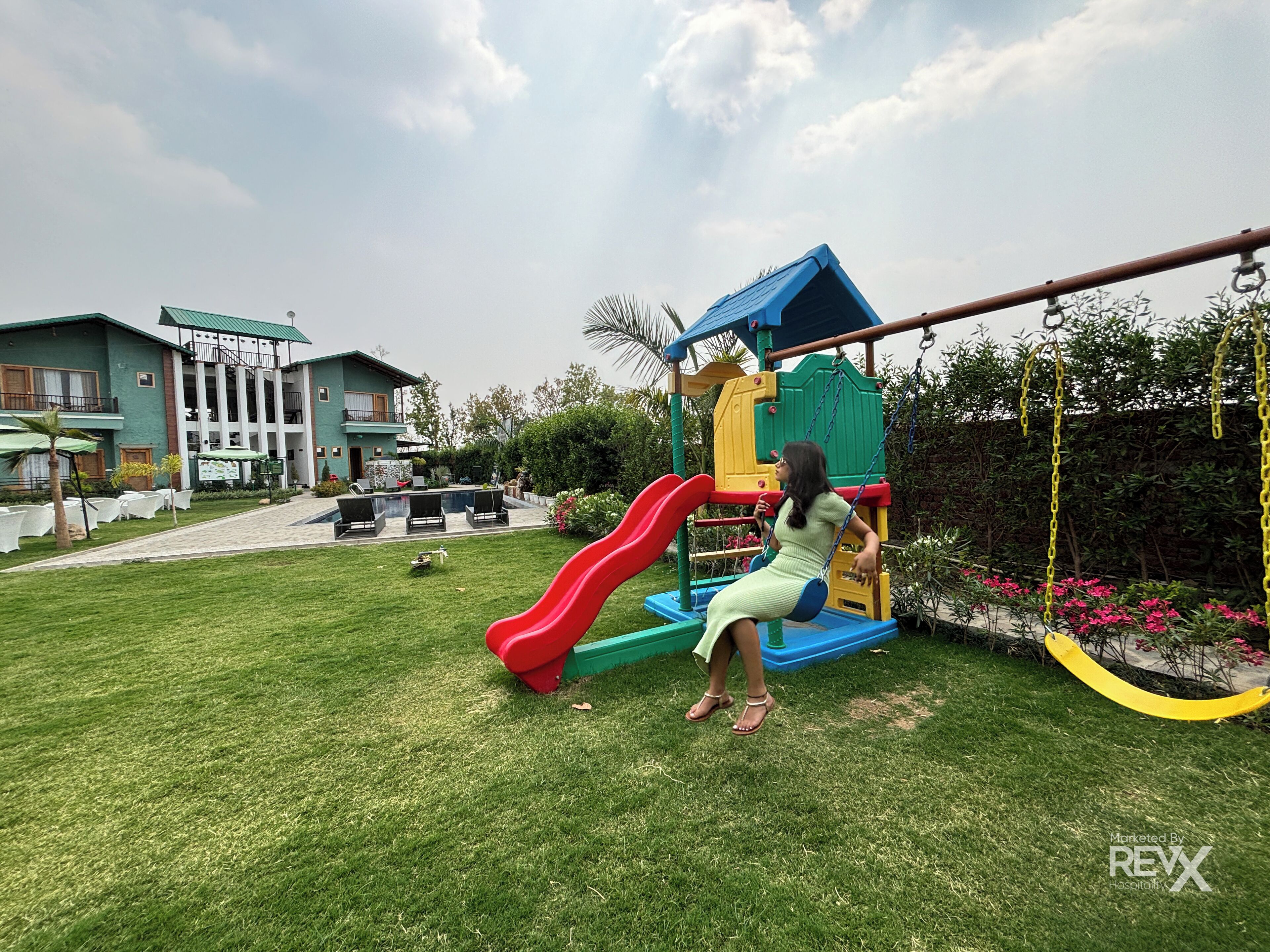 A playground with a slide and swings, surrounded by a well-maintained lawn and a building with a balcony.